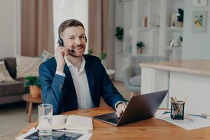 lachende zakenman in koptelefoon zittend aan een bureau op kantoor aan huis en werken op laptop foto