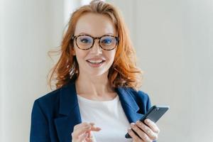 headshot van vrouwelijke bankier met aangenaam uiterlijk en rood haar, bankiert online op mobiele telefoon, controleert melding op financiële website, draagt transparante bril en elegant formeel pak foto