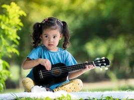 Aziatisch klein meisje zittend op het tapijt, ontspannen en ukelele spelen buiten school om te genieten in het natuurpark foto