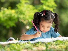 een Aziatisch meisje ligt op het tapijt en beschilderd met kleurpotloden, wat leuk is om buiten de school in het natuurpark te leren foto