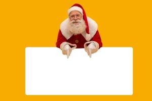 gelukkige kerstman die uitkijkt van achter het lege bord geïsoleerd op een gele achtergrond met kopieerruimte foto