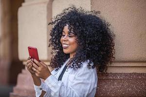 jonge krullend haar zwarte vrouw lopen met behulp van mobiele telefoon. sms'en op straat. grote stad. foto