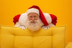 Kerstman zittend op een gele bank op gele achtergrond met kopieerruimte. gele bank. foto