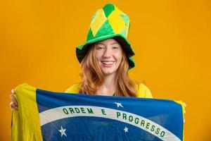 brazilie aanhanger. braziliaanse roodharige vrouw fan vieren op voetbal, voetbalwedstrijd op gele achtergrond. braziliaanse kleuren. het dragen van een t-shirt, vlag en waaierhoed. foto