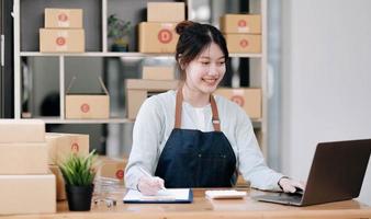een portret van een jonge aziatische vrouw e-commerce werknemer zittend in het kantoor vol pakketten op de achtergrond notitie van bestellingen en een rekenmachine, voor mkb-bedrijven e-commerce en bezorging bedrijf. foto