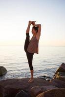 fitness gemengd ras aziatische vrouw in yoga pose op het ochtendstrand, mooie fit vrouw praktijk fitness exrxise stenen, ochtend zee of oceaan achtergrond foto