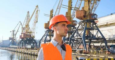 mannelijke arbeider van zeehaven in oranje helm en veiligheidswest, kranen en zeeachtergrond foto