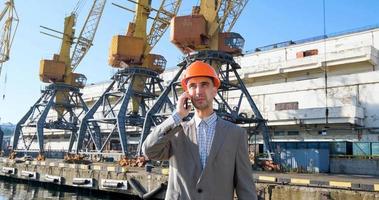 jonge mannelijke arbeider van zeehaven in helm, vrachtmanager in pak en halmet werkt buiten, kranen en zee-achtergrond foto