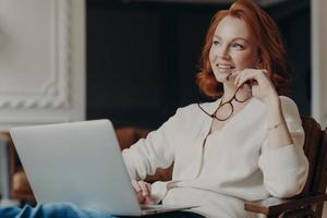 horizontaal schot van nadenkende jonge roodharige vrouw ergens geconcentreerd, werkt aan freelance project, houdt een bril vast, houdt laptop op knieën, geniet van online baan, creëert nieuwe publicatie foto