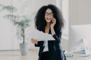 tevreden vrouwelijke directeur met krullend haar gekleed in formele outfit, houdt documenten vast, belt via mobiele telefoon, staat in de buurt van werkplek met computer, deelt mening over opstarten, maakt bedrijfsrapport foto