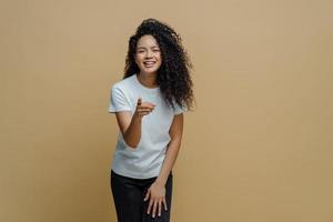 vrolijke energieke jonge vrouw met krullend afrohaar wijst wijsvinger naar voren op camera, voelt zich positief en kiest iemand, draagt wit t-shirt en spijkerbroek, ziet iets leuks vooraan. foto