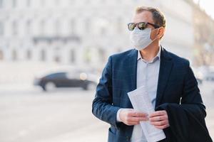 covid19. man werknemer gekleed in formele kleding, houdt krant in handen, poseert op straat, draagt chirurgisch masker, vermijdt drukke straat om geen virus te krijgen, poseert tegen onscherpe achtergrond met transport foto