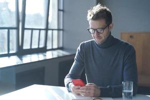 knappe Duitse zakenman in brillen zittend aan een bureau op kantoor aan huis en het gebruik van smartphone foto