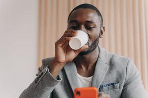 elegante Afrikaanse etniciteit zakenman die afhaalkoffie drinkt terwijl hij op het scherm van de mobiele telefoon kijkt foto