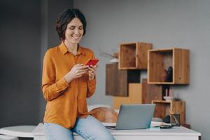 jonge gelukkige europese vrouw freelancer zittend op tafel op kantoor aan huis en het gebruik van mobiele telefoon, foto