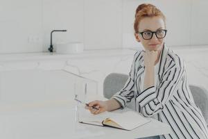 nadenkende roodharige vrouw afgeleid van het werk maakt aantekeningen in notitieblok houdt pen vast foto