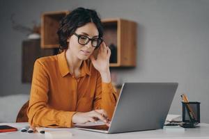 ocused Spaanse vrouw met een bril zit aan het bureau en typt op laptop tijdens thuiswerk op afstand foto