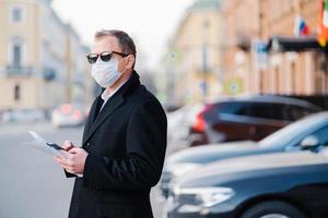 pandemische uitbraak van het coronavirus. serieuze zakenman poseert buiten in de buurt van vervoer op straat, houdt moderne mobiele telefoons en kranten vast, gekleed in zwarte jas, draagt beschermend masker tegen coronavirus. foto