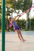 mooie fitte vrouw in roze en paarse sportkleding training op buitengymnastiek in de ochtend, oefeningen met ophangriemen in park foto