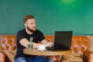jonge, bebaarde roodharige man werkt in het café met mobiele telefoon en laptop