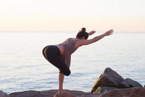 fitness gemengd ras aziatische vrouw in yoga pose op het ochtendstrand, mooie fit vrouw praktijk fitness exrxise stenen, ochtend zee of oceaan achtergrond foto