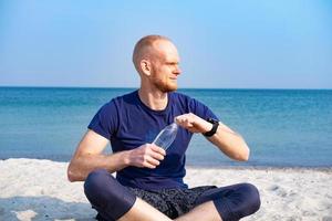 jonge mannelijke atleet die vers water uit een plastic fles op het strand haalt foto