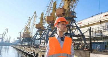 mannelijke arbeider van zeehaven in oranje helm en veiligheidswest, kranen en zeeachtergrond foto