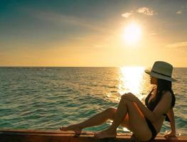 sexy en ontspannen vrouw dragen zwarte bikini met stro hoed zitten op houten balk in de buurt van zandstrand bij zonsondergang. meisje geniet van vakantie op tropisch paradijsstrand op zomervakantie. vakantie reizen. zomerse sferen. foto