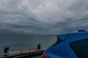 selectieve focus op kofferbak van blauwe luxe suv-auto geparkeerd op betonnen weg naast zeestrand op regenachtige dag met donkere stormhemel en oceaanwater. regendruppel op blauwe suv-auto. roadtrip reizen. natte parkeerplaats. foto