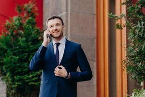 portret van knappe tevreden succesvolle zakenman praat op mobiele telefoon, bespreekt banktransacties, gekleed in pak, heeft positieve uitdrukking. bedrijfs- en technologieconcept foto