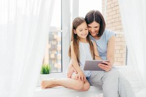 horizontaal schot van mooie brunette moeder en dochter brengen samen vrije tijd door, zitten op de vensterbank, kijken naar interessante film via digitale tablet, in de slaapkamer zijn. mensen, technologie, familieconcept foto