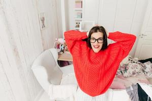 bovenaanzicht van blauwe ogen, brunette jonge vrouw in casual rode trui, draagt rechthoekige bril, staat in haar ruime kamer thuis, glimlacht graag. mensen, schoonheid en lifestyle concept. foto