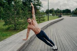 atletische man staat in de zijplankhouding, heft een arm op, poseert met een shirtloze torso, draagt een broek en sluipt. sportman oefent in het park op frisse lucht, heeft een perfect fit spierenlichaam. gezonde levensstijl foto