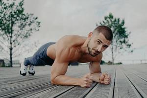 motiverende bebaarde man staat in plank pose, traint spieren en heeft een sterk lichaam. sportieve volwassen man voert push-up oefening buiten uit. bodybuilder heeft buiktraining. actief levensstijlconcept foto