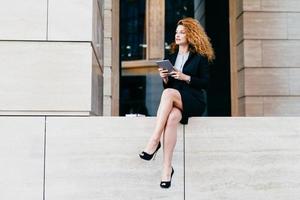 slanke zakenvrouw met golvend luxueus haar, met slanke benen, gekleed in een zwart elegant pak en schoenen, met een tabletcomputer, opzij kijkend terwijl ze een minuut rust na hard werken op kantoor foto