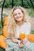 verticaal schot van een knappe, mooie jonge blonde vrouw met krullend haar, positieve glimlach, brengt vrije tijd door met vrienden, heeft samen een feestje, poseert in een hangende stoel, drinkt een frisse zomercocktail. foto