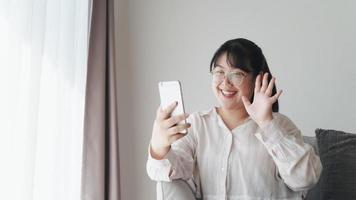 jonge aziatische vrouw die smartphone gebruikt voor online videoconferentiegesprek zwaaiende hand hallo gebaar maken op de bank in de woonkamer. foto