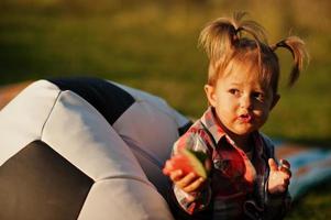 schattig babymeisje in geruit overhemd zit op de poef van de voetbalbal en eet watermeloen. foto