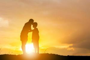 silhouet van een moeder die zijn zoon kust in een voorhoofd op de zonsondergang zomerdag. foto