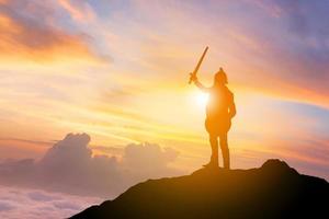 silhouet van zakenman ridder zwaard en schild op bergtop zonsondergang achtergrond, strijd zakelijke krijger concept. foto