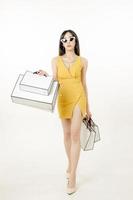 zelfverzekerde aziatische vrouw in een zwoele gele jurk die een zonnebril draagt en een boodschappentas draagt, heeft plezier bij het winkelen. foto
