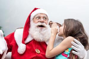 klein meisje fluistert in het oor van de kerstman. een geheim vertellen. het onthullen van het geschenk dat u zou willen winnen. praten over hoe ze zich gedurende het jaar gedroeg. foto