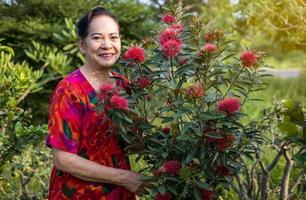 het portret van een Thaise oudere vrouw die staat en lacht met bloemen in een tuin. foto