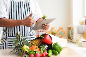 close-up van de hand van de chef die tablet gebruikt om online groenten en fruit te kopen. en er zijn groenten en fruit en papieren zakken op de keukentafel. online winkelen en bezorgconcept. foto