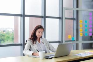 portret van geconcentreerde en zelfverzekerde blanke zakenvrouw werkt, schrijft het plan en de informatie in een notitieblok en gebruikt een laptop op het bureau. start-up moderne kantoor of werkplek achtergrond. foto