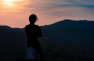 achteraanzicht van een vrouw die op een hek zit en 's ochtends naar de zonsopgang boven de berg kijkt. avonturen reis. vrouw reist alleen. mensen met een rustige geest van nature. zitten om aan het verleden te denken. foto