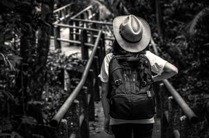 zwart-wit foto van aziatische vrouw toerist met hoed en rugzak staande en beginnen te lopen op natuurpad brug in groenblijvende bos. alleen jonge vrouw reiziger.