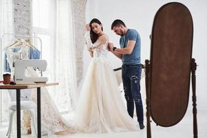 man helpt meisje terwijl ze in de spiegel kijkt. het proces van het passen van de jurk in de studio van handgemaakte kleding foto
