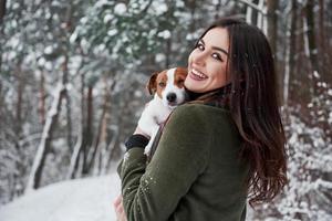 meisje is haar hoofd naar de camera gedraaid. lachende brunette die plezier heeft tijdens het wandelen met haar hond in het winterpark foto