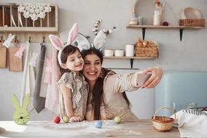 met behulp van wit gekleurde smartphone. moeder en dochter in konijnenoren met Pasen hebben overdag plezier in de keuken foto